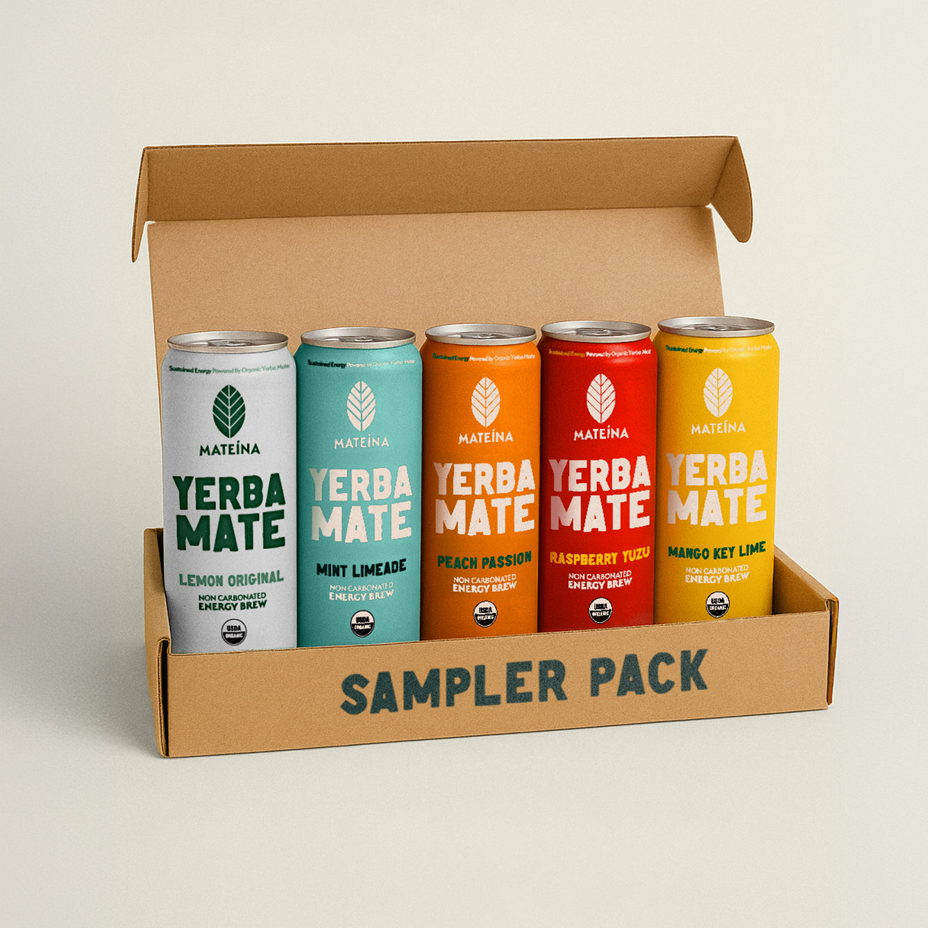 An open cardboard box labeled ‘Sampler Pack’ containing five cans of Mateína Yerba Mate. The cans are arranged side by side and feature different flavors: Lemon Original (white can), Mint Limeade (light green can), Peach Passion (orange can), Raspberry Yuzu (red can), and Mango Key Lime (yellow can). Each can is labeled as a non-carbonated energy brew made with organic yerba mate.
